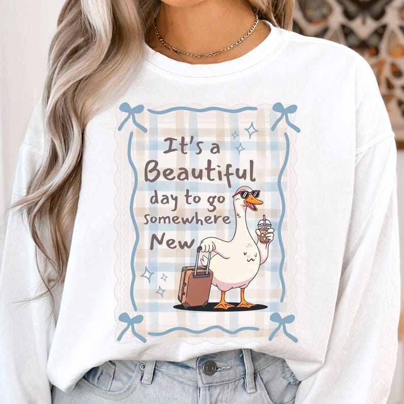 May include: White long-sleeved sweatshirt with a graphic of a goose wearing sunglasses and holding a drink and suitcase. The text reads "It's a Beautiful day to go somewhere New."