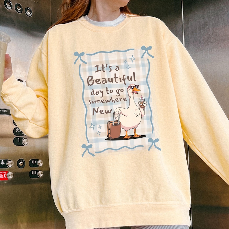 May include: A light yellow sweatshirt featuring a cartoon goose with sunglasses, a suitcase, and a drink. The goose is inside a blue plaid frame with the text "It's a Beautiful day to go Somewhere New."