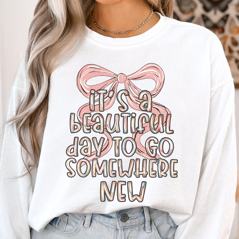 Puede incluir: Sudadera blanca de manga larga con un lazo rosa y el texto "It's a beautiful day to go somewhere new" en una fuente colorida y juguetona. Un estilo casual y c&oacute;modo.