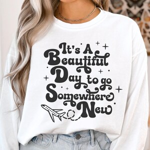 以下が含まれることがあります： 「It's A Beautiful Day to go Somewhere New」の黒い文字がプリントされた白いスウェットシャツ。小さな飛行機のグラフィックと星のデザイン。長袖クルーネックのスウェットシャツです。