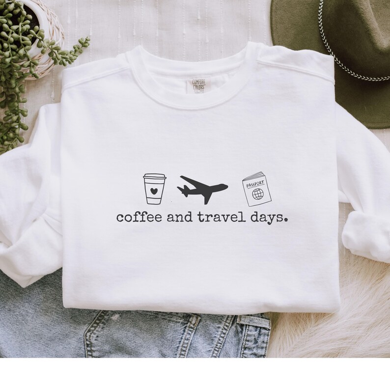 Puede incluir: Sudadera blanca con un gr&aacute;fico negro de una taza de caf&eacute;, un avi&oacute;n y un pasaporte. El texto "coffee and travel days" est&aacute; impreso debajo. La sudadera est&aacute; hecha de un material suave.