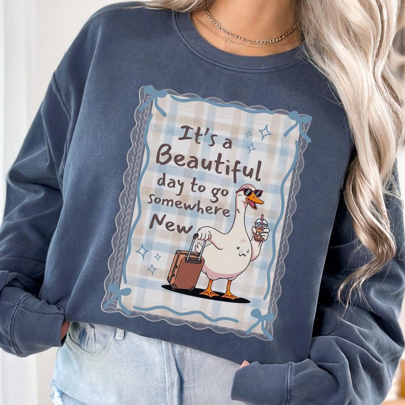 May include: A navy blue sweatshirt features a graphic of a goose wearing sunglasses and holding a coffee cup and suitcase. The graphic is framed by a decorative border and includes the text "It's a Beautiful day to go Somewhere New."