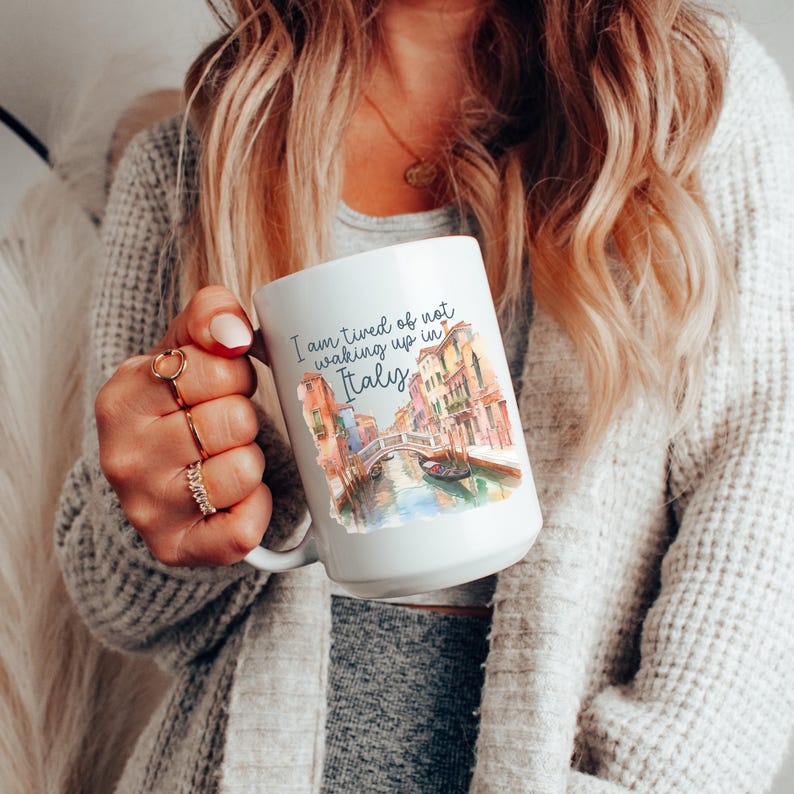 May include: White ceramic coffee mug with a watercolor illustration of a canal in Venice, Italy. The text on the mug reads "I am tired of not waking up in Italy."
