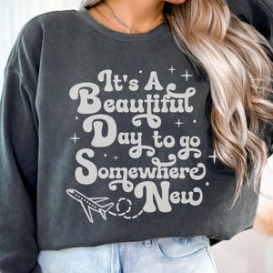 以下が含まれることがあります： 「It's A Beautiful Day to go Somewhere New」の白文字と飛行機のグラフィックが特徴の濃いグレーのスウェットシャツ。クルーネックのスウェットシャツは長袖です。