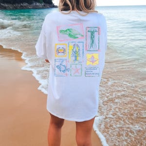 May include: White t-shirt with colorful illustrations of sea creatures, including a turtle, crab, lobster, fish, starfish, jellyfish, and stingray. The text "Saltwater Heals Everything" is printed below the illustrations. The shirt is worn on a beach.