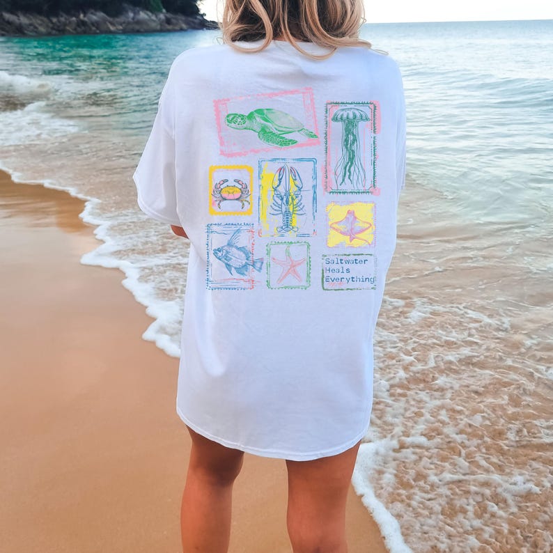 May include: White t-shirt with colorful illustrations of sea creatures, including a turtle, crab, lobster, fish, starfish, jellyfish, and stingray. The text "Saltwater Heals Everything" is printed below the illustrations. The shirt is worn on a beach.
