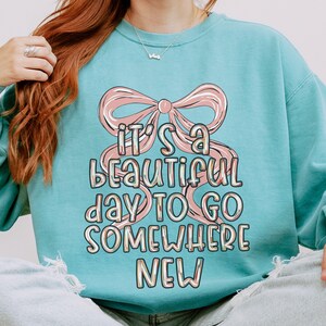 Puede incluir: Una sudadera azul claro con un lazo rosa y el texto "IT'S A BEAUTIFUL DAY TO GO SOMEWHERE NEW". El dise&ntilde;o tiene una est&eacute;tica retro y pastel. La sudadera es de estilo casual y c&oacute;modo.
