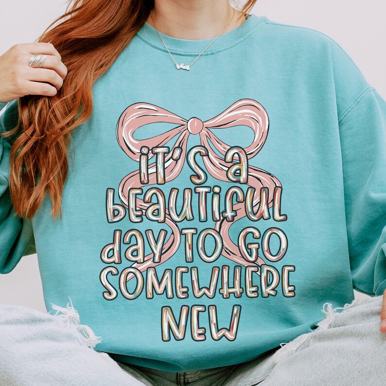 Puede incluir: Una sudadera azul claro con un lazo rosa y el texto "IT'S A BEAUTIFUL DAY TO GO SOMEWHERE NEW". El dise&ntilde;o tiene una est&eacute;tica retro y pastel. La sudadera es de estilo casual y c&oacute;modo.