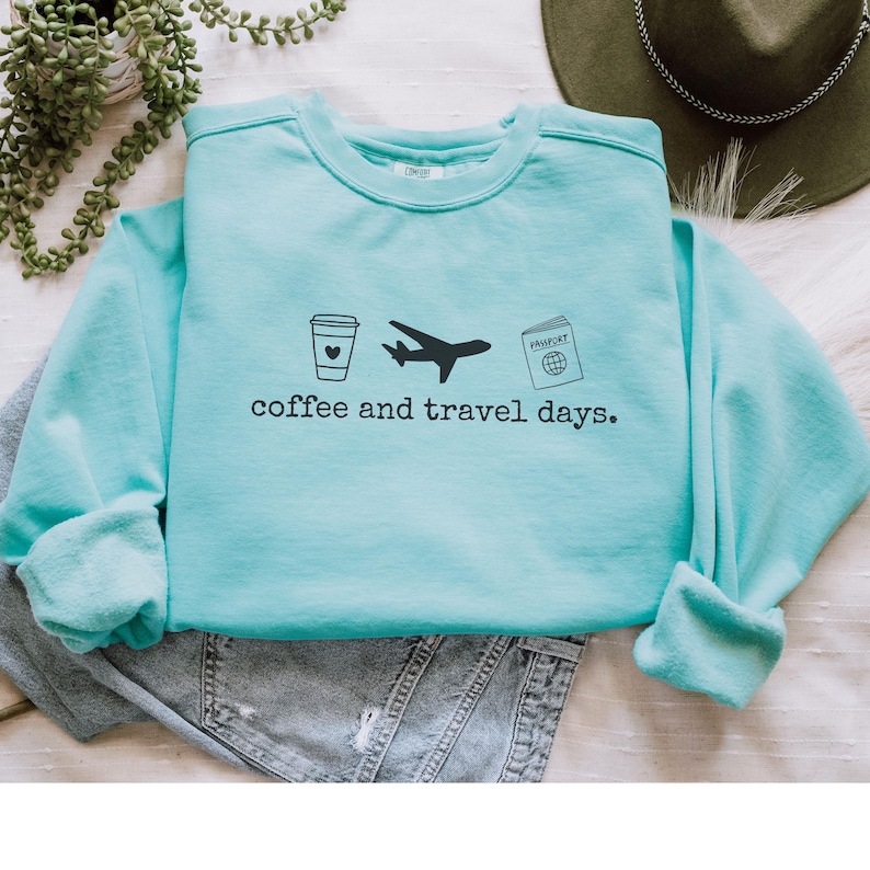 Puede incluir: Sudadera azul claro con la frase "coffee and travel days" en negro. El dise&ntilde;o incluye una taza de caf&eacute;, un avi&oacute;n y un pasaporte. La sudadera est&aacute; doblada sobre unos vaqueros grises.