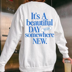 K&ouml;nnte beinhalten: Wei&szlig;es Sweatshirt mit blauem Text: "It's A beautiful DAY to go somewhere NEW." Das Sweatshirt hat lange &Auml;rmel und einen Rundhalsausschnitt. Der Text ist fett und ohne Serifen.