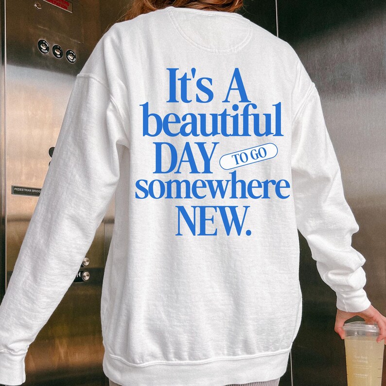 K&ouml;nnte beinhalten: Wei&szlig;es Sweatshirt mit blauem Text: "It's A beautiful DAY to go somewhere NEW." Das Sweatshirt hat lange &Auml;rmel und einen Rundhalsausschnitt. Der Text ist fett und ohne Serifen.