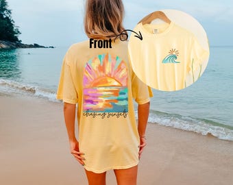 May include: A light yellow t-shirt with a colorful sunset graphic on the back and a small wave and sun design on the front. The back graphic features a vibrant sunset over water with the words "chasing sunsets".
