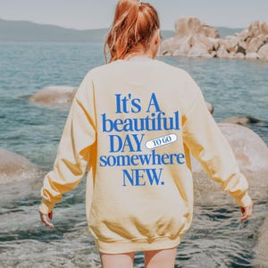 K&ouml;nnte beinhalten: Ein hellgelbes Sweatshirt mit dem Text "It's A beautiful DAY to go somewhere NEW" in Blau. Das Sweatshirt hat einen Rundhalsausschnitt und lange &Auml;rmel. Der Hintergrund zeigt ein Gew&auml;sser und Felsen.