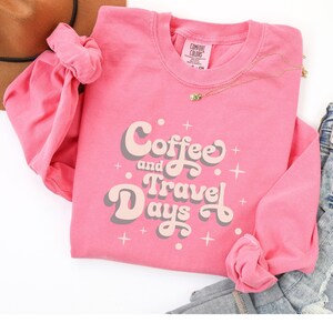 May include: A pink long-sleeved sweatshirt with the words "Coffee and Travel Days" in a retro font. The shirt has a crew neck and is adorned with small cross symbols. A gold necklace and a scrunchie are also visible.