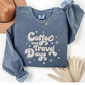 May include: A blue crewneck sweatshirt with the words "Coffee and Travel Days" in a retro font. The sweatshirt has a faded appearance and a small tag on the neckline. A gold necklace and a brown hat are visible.