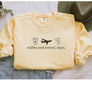 Puede incluir: Sudadera amarilla clara con la frase "coffee and travel days" impresa debajo de un gr&aacute;fico de taza de caf&eacute;, avi&oacute;n y pasaporte. La sudadera es de cuello redondo.