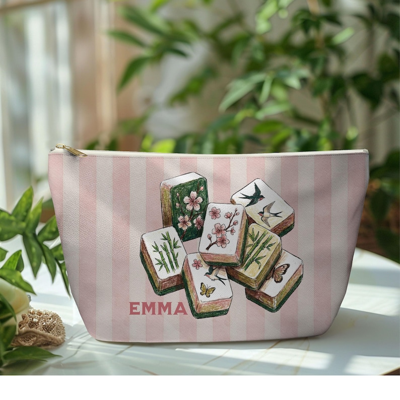 May include: A pink and white striped cosmetic bag with illustrated Mahjong tiles. The tiles show floral, bamboo, and bird designs. The name "EMMA" is printed in pink on the bag.