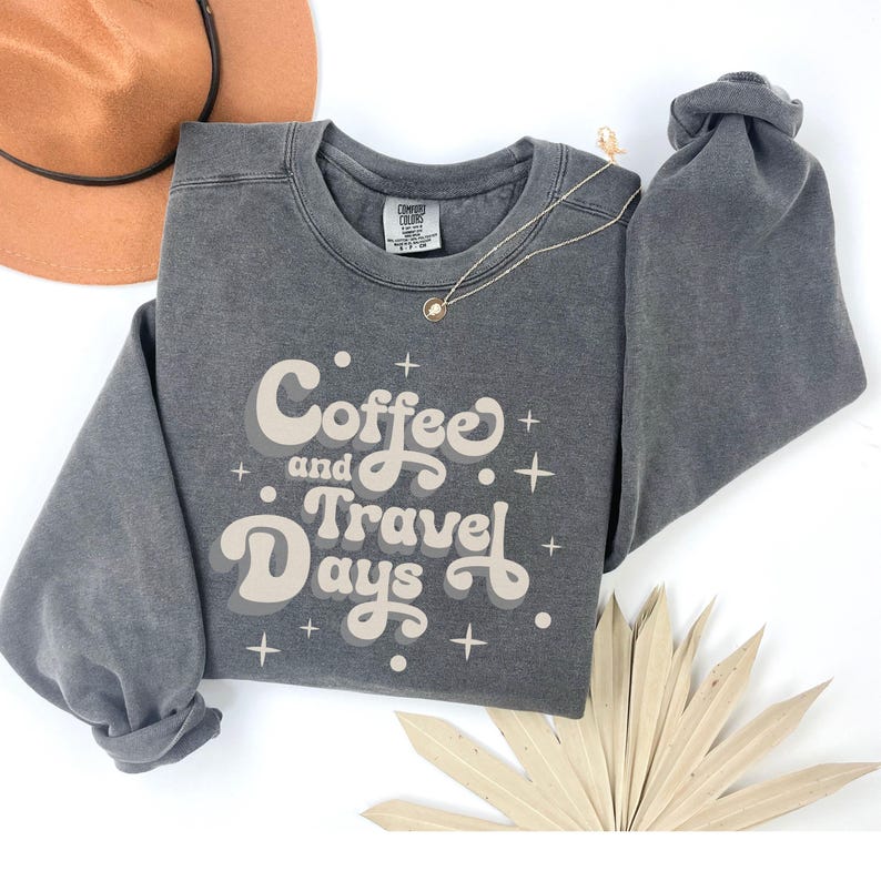 May include: A charcoal gray sweatshirt with the words "Coffee and Travel Days" in a retro font. The sweatshirt has a crew neck and long sleeves. A gold necklace and a brown hat are also visible.