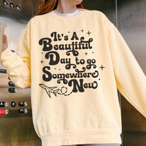 以下が含まれることがあります： 「It's A Beautiful Day to go Somewhere New」の黒い文字が入った、薄い黄色のスウェットシャツ。テキストの下には小さな飛行機のグラフィックがあります。スウェットシャツは長袖です。