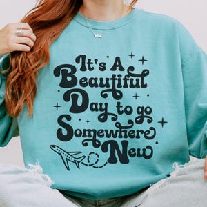 以下が含まれることがあります： 黒い文字で「It's A Beautiful Day to go Somewhere New」というフレーズがプリントされた、ターコイズ色のスウェットシャツ。テキストの下には小さな飛行機のグラフィックがあります。スウェットシャツはクルーネックと長袖です。