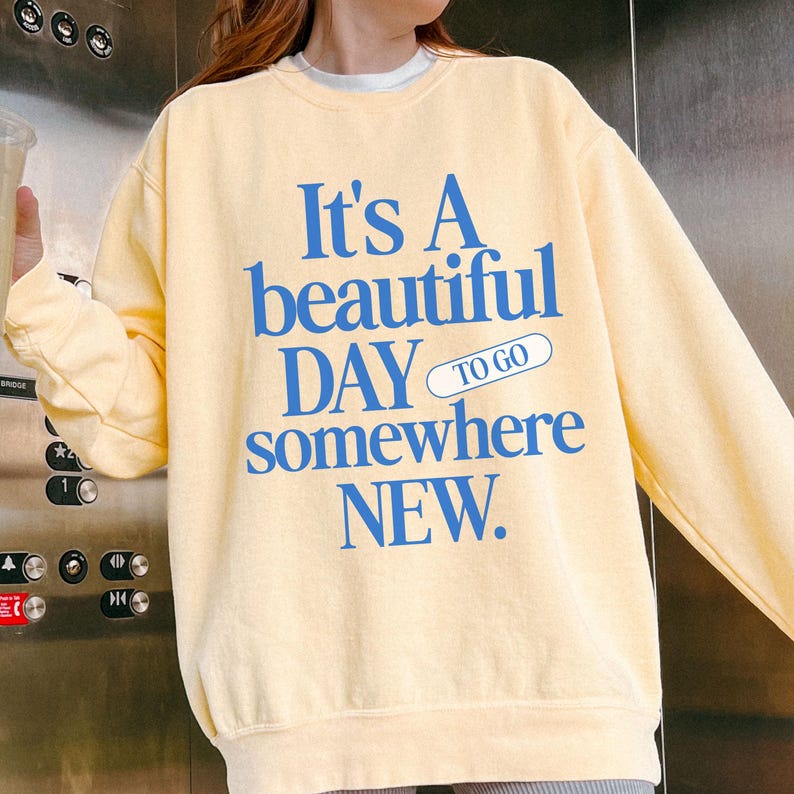 K&ouml;nnte beinhalten: Hellgelbes Sweatshirt mit blauem Text: "It's A beautiful DAY to go somewhere NEW". Das Sweatshirt hat lange &Auml;rmel und einen Rundhalsausschnitt. Das Design ist mittig auf der Vorderseite platziert.