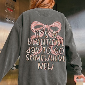Puede incluir: Sudadera gris oscuro con un lazo rosa y el texto "IT'S A BEAUTIFUL DAY TO GO SOMEWHERE NEW" en una fuente colorida y juguetona. La sudadera tiene mangas largas y cuello redondo.