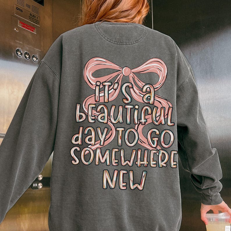 Puede incluir: Sudadera gris oscuro con un lazo rosa y el texto "IT'S A BEAUTIFUL DAY TO GO SOMEWHERE NEW" en una fuente colorida y juguetona. La sudadera tiene mangas largas y cuello redondo.