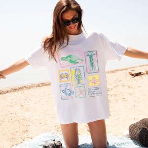 May include: White t-shirt featuring colorful illustrations of sea creatures, including a turtle, jellyfish, crab, and fish. The shirt also has the text "Saltwater Heals Everything". The shirt is worn on a sunny beach.