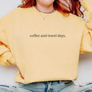 May include: A light yellow sweatshirt with the text "coffee and travel days." in a simple, black font. The sweatshirt is a solid color and appears to be made of a soft material. The person wearing the sweatshirt is also wearing a gold necklace.