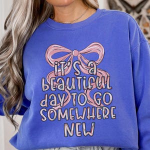 Puede incluir: Sudadera azul b&iacute;garo con la frase "IT'S A BEAUTIFUL DAY TO GO SOMEWHERE NEW" en una fuente caprichosa. El texto est&aacute; superpuesto con un lazo rosa.