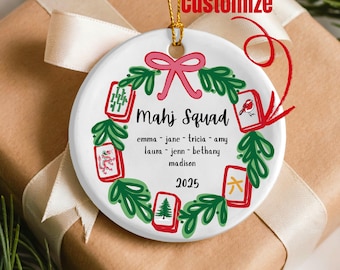 May include: A white ceramic ornament with a gold string for hanging. The ornament features the text "Mahj Squad" and a list of names. A wreath of green leaves and red Mahjong tiles surrounds the text. A pink bow is at the top. The year 2025 is at the bottom.