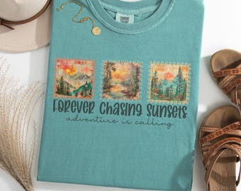 May include: Teal-colored t-shirt with three landscape images and the text "Forever Chasing Sunsets, adventure is calling." The shirt is styled with a gold necklace, a beige hat, and brown sandals.