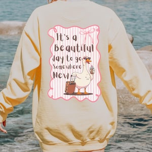 Puede incluir: Sudadera de color crema con un gr&aacute;fico de un ganso con gafas de sol, sosteniendo una bebida y una maleta. El gr&aacute;fico est&aacute; enmarcado por un borde rosa e incluye el texto "It's a Beautiful day to go Somewhere New."
