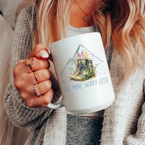 May include: White ceramic mug with a watercolor illustration of hiking boots in a mountain landscape. The text "Hike More Worry Less" is printed in blue below the illustration.