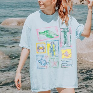 May include: Light blue t-shirt featuring colorful illustrations of sea creatures, including a turtle, jellyfish, and crab, within decorative frames. The shirt also has the text "Saltwater Heals Everything."