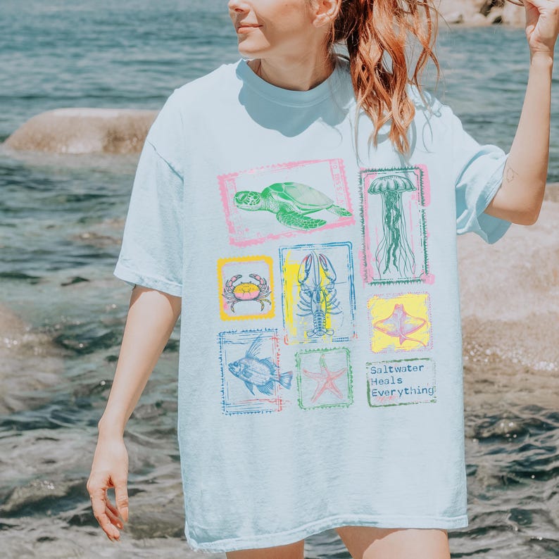 May include: Light blue t-shirt featuring colorful illustrations of sea creatures, including a turtle, jellyfish, and crab, within decorative frames. The shirt also has the text "Saltwater Heals Everything."