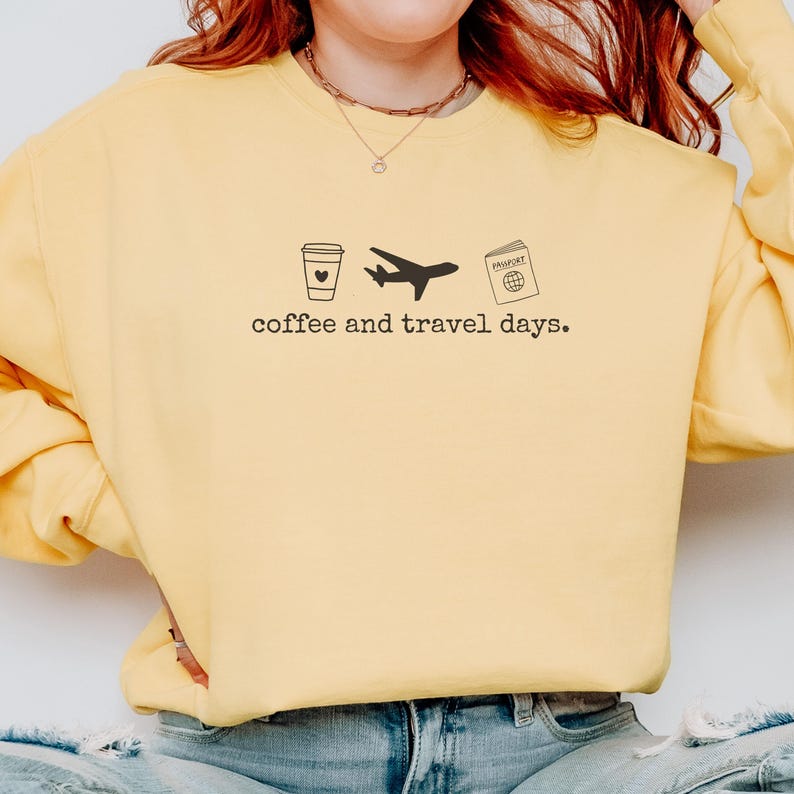 Puede incluir: Sudadera amarilla con un gr&aacute;fico negro de una taza de caf&eacute;, un avi&oacute;n y un pasaporte. El texto "coffee and travel days." est&aacute; impreso debajo. La sudadera es de estilo casual.