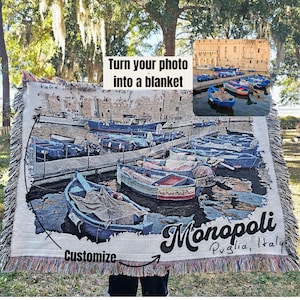 May include: A woven tapestry blanket featuring a photo of colorful boats docked in a harbor. The blanket has a white background with a fringe border. The text "Turn your photo into a blanket" and "Monopoli Puglia, Italy" is printed on the blanket.
