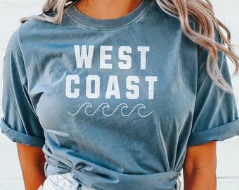 May include: A blue t-shirt with the text "WEST COAST" in white lettering and a row of white waves underneath. The shirt is tied at the waist.