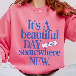 K&ouml;nnte beinhalten: Rosa Sweatshirt mit blauem Text: "It's A beautiful DAY to go somewhere NEW." Das Wort "to go" befindet sich in einem wei&szlig;en Pillen-Design. Das Sweatshirt hat eine einfarbige rosa Farbe.