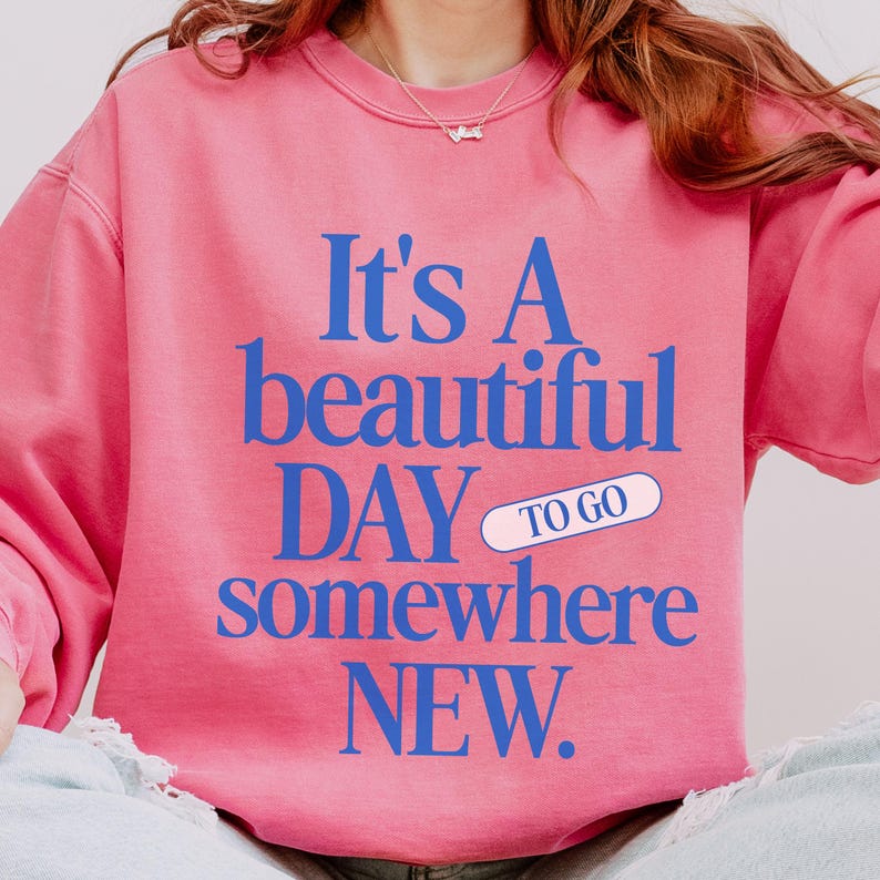 K&ouml;nnte beinhalten: Rosa Sweatshirt mit blauem Text: "It's A beautiful DAY to go somewhere NEW." Das Wort "to go" befindet sich in einem wei&szlig;en Pillen-Design. Das Sweatshirt hat eine einfarbige rosa Farbe.