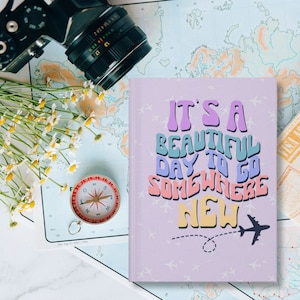 May include: A purple notebook with the text "IT'S A BEAUTIFUL DAY TO GO SOMEWHERE NEW" in colorful, retro-style lettering. The notebook has a plane and a circle with an arrow pointing to the right.