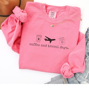 Puede incluir: Una sudadera rosa con la frase "coffee and travel days" impresa en negro. El dise&ntilde;o incluye una taza de caf&eacute;, un avi&oacute;n y un pasaporte. Tambi&eacute;n se ven un collar dorado y unos vaqueros.