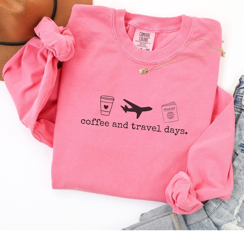 Puede incluir: Una sudadera rosa con la frase "coffee and travel days" impresa en negro. El dise&ntilde;o incluye una taza de caf&eacute;, un avi&oacute;n y un pasaporte. Tambi&eacute;n se ven un collar dorado y unos vaqueros.