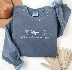 Puede incluir: Una sudadera azul gris&aacute;ceo con las palabras "coffee and travel days" impresas en blanco. El gr&aacute;fico incluye una taza de caf&eacute;, un avi&oacute;n y un pasaporte. Un collar dorado est&aacute; drapeado sobre la sudadera.