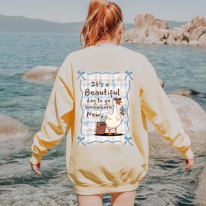 May include: A light yellow sweatshirt with a graphic on the back. The graphic features a cartoon goose with sunglasses, a suitcase, and a coffee cup, with the text "It's a Beautiful day to go Somewhere New."