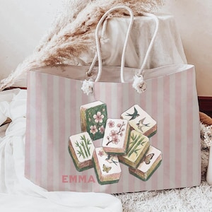 May include: A large tote bag with pink and white vertical stripes. The bag features a design of colourful Mahjong tiles with floral and bird illustrations. The name "EMMA" is printed in pink on the front. The bag has white rope handles.