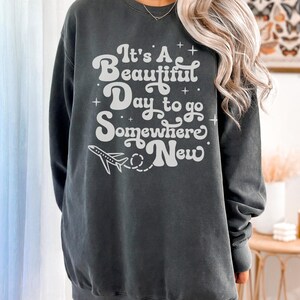 以下が含まれることがあります： 「It's A Beautiful Day to go Somewhere New」の白文字と飛行機のグラフィックが特徴のダークグレーのスウェットシャツ。クルーネックのスウェットシャツはカジュアルなスタイルです。