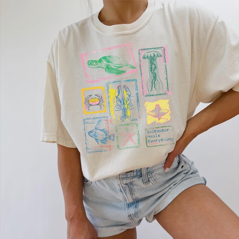 May include: Cream-colored t-shirt featuring colorful sea creatures in square frames. Designs include a turtle, jellyfish, crab, lobster, fish, starfish, and a stingray. The text "Saltwater Heals Everything" is printed on the shirt.