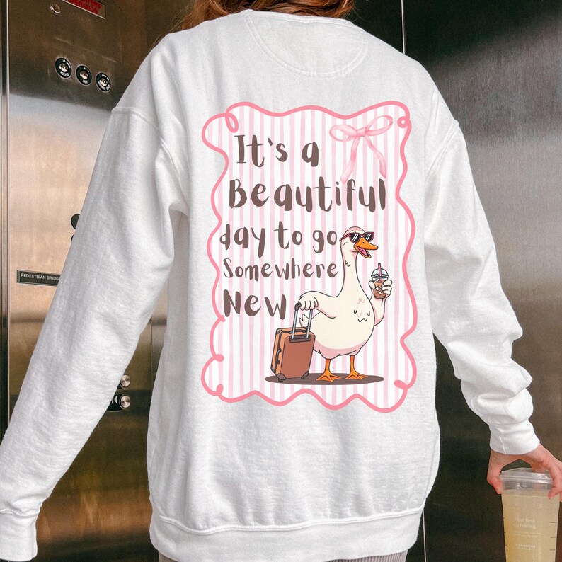 Puede incluir: Sudadera blanca con un dise&ntilde;o de rayas rosas y blancas con un ganso de dibujos animados con gafas de sol y sosteniendo una bebida y una maleta. El texto de la sudadera dice "It's a Beautiful day to go Somewhere New."
